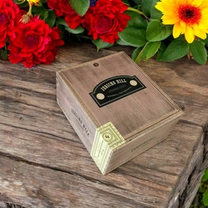 May include: A wooden box with the text "JERICHO HILL" and "ESTELI, NICARAGUA" on the lid. The box is light brown with a distressed finish and a small label on the corner. Red and yellow flowers are in the background.
