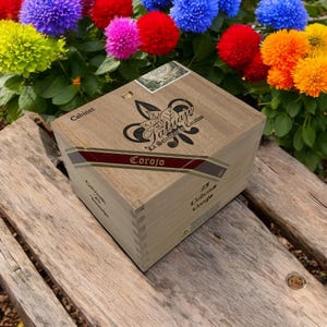 May include: A wooden box with the text "Tatuaje La Seleccion de Cazador Corojo" and a leaf design. The box is sitting on a wooden surface with colorful flowers in the background.