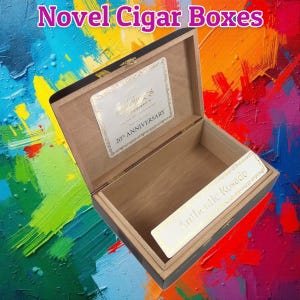 May include: Open wooden cigar box with a black exterior and a light brown interior. The box has a gold clasp and two white labels with gold text that read "Sublimes 20th Anniversary" and "Authentic Rosado". The text "Novel Cigar Boxes" is at the top.