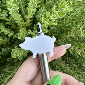 May include: A white plastic pig figurine attached to a silver metal pen. The pen is held in a hand with green nail polish.