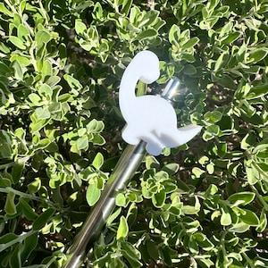 May include: A white dinosaur-shaped straw topper attached to a silver metal straw. The straw and topper are lying on a bed of green leafy plants.