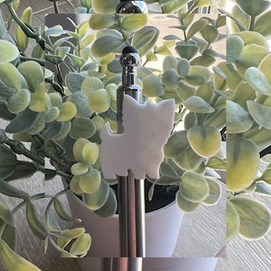 May include: A silver stylus with a black tip and a white dog-shaped charm. The stylus is set against a backdrop of green leafy plants in a white pot, creating a clean and modern aesthetic.