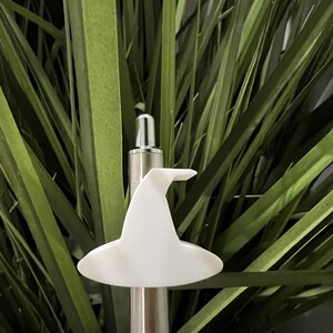 May include: A white witch hat-shaped bottle stopper. The hat is made of a smooth, opaque material and is mounted on a silver-coloured metal rod. The background features green foliage.