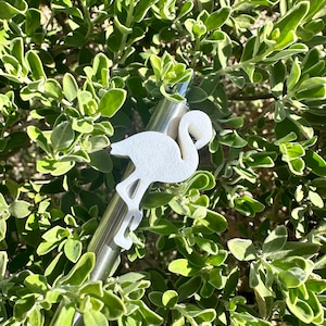 May include: A stainless steel reusable straw with a white flamingo-shaped topper. The straw is lying on a bed of green leaves.