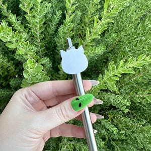May include: A silver pen with a white cat head topper. The pen is being held by a hand with green alien nail art.