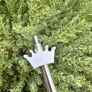 May include: A white crown-shaped bottle stopper on a silver metal stem. The stopper is set against a backdrop of green and white leafy foliage. The crown design adds a touch of elegance.