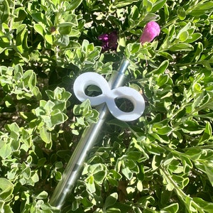 May include: A stainless steel straw with a white infinity symbol-shaped topper. The straw is set against a backdrop of green foliage with small purple flowers, creating a natural and vibrant contrast.