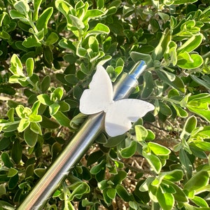 May include: A silver metal pen with a white butterfly charm attached to the top. The pen is lying on a bed of green leaves.