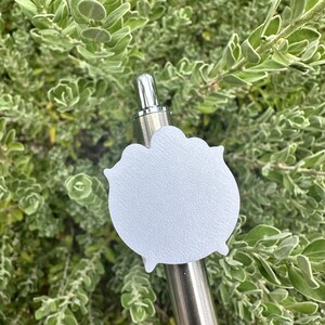 May include: A silver metal pen with a white, cloud-shaped charm attached. The charm has a textured surface and is set against a backdrop of green foliage. The pen's design is simple and elegant.