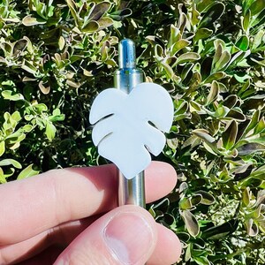 May include: A silver pen with a white, heart-shaped monstera leaf charm. The pen is held in a hand against a background of green foliage.