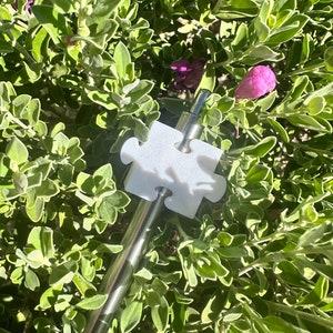 May include: A white puzzle piece charm attached to a silver metal straw. The straw and charm are lying on top of green foliage.