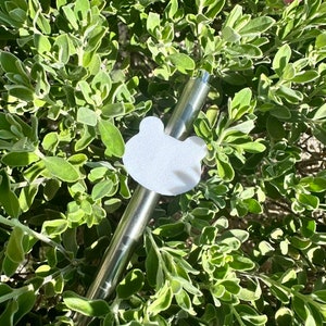 May include: A silver reusable straw with a white frog-shaped charm attached to it. The straw is lying on a bed of green leaves.