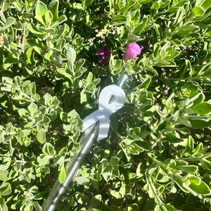 May include: A stainless steel straw with a white ribbon-shaped attachment, set against a backdrop of green foliage and small purple flowers. The straw is angled diagonally, creating a contrast with the natural background.