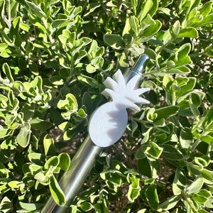 May include: A silver pen with a white pineapple-shaped topper. The pen is lying on a bed of green leaves.