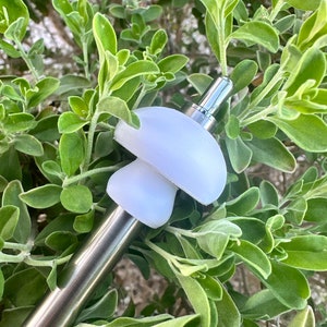 May include: A stainless steel and white mushroom-shaped bottle stopper. The stopper is set against a backdrop of green foliage. The design features a mushroom cap and stem, with a sleek, modern aesthetic.