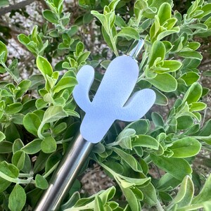May include: A silver pen with a light blue cactus-shaped topper. The pen is lying on a bed of green leaves.