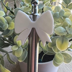 May include: A white bow-shaped stylus attachment on a silver stylus. The stylus has a black tip and is set against a backdrop of green leafy plants in a white pot. The bow is made of a smooth, solid material.