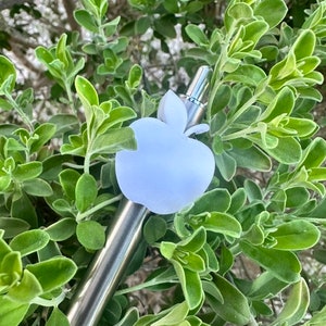 May include: A silver metal straw with a white apple-shaped silicone tip. The straw is partially visible against a background of green foliage.