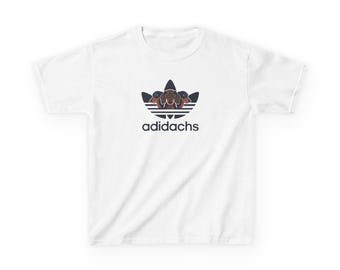 Può includere: T-shirt bianca con un logo parodia con un design di cane bassotto. Il logo è una parodia del logo Adidas, con la parola "adidachs" in nero, carattere sans-serif sotto. La maglietta è realizzata in un materiale morbido e confortevole.