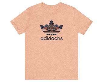 Può includere: Una t-shirt a maniche corte color pesca mélange con una grafica blu scuro di tre teste di bassotto disposte a forma di trifoglio, con la scritta adidachs stampata in lettere blu scuro direttamente sotto il disegno.