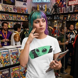 May include: A white short-sleeved t-shirt featuring a graphic print of a green alien face with a raised hand inside a black horizontal brushstroke shape. The alien has large dark eyes and a smooth head, centred on the chest of the garment.