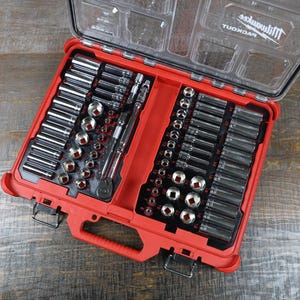 以下が含まれることがあります： 赤と透明なプラスチック製のツールボックス。さまざまなシルバーのソケットとラチェットレンチが入っています。ツールボックスは開いており、ツールが整理されて配置されています。「Milwaukee PACKOUT」のブランド名が蓋に表示されています。