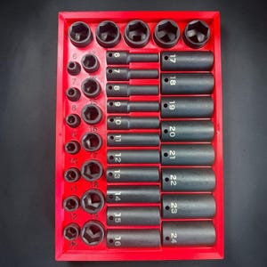 May include: A red plastic organizer tray containing a comprehensive set of black metal impact sockets. The tray features labeled slots for various sizes, including small and large sockets ranging from 6 to 24, organized in rows and columns.