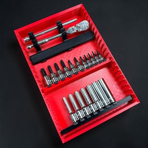 May include: A red tool organiser tray containing various chrome sockets, extensions, and a ratchet wrench. The tools are neatly arranged in black holders within the tray. The tray is on a black surface.