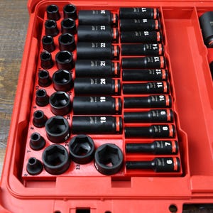 May include: A red toolbox with a clear lid, filled with a set of black impact sockets. The sockets are arranged in rows and labeled with sizes from 6 to 24. The toolbox has a metal latch.