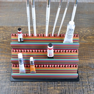 Puede incluir: Un organizador de suministros de arte a rayas multicolores que contiene pinceles y tubos de pintura. El organizador tiene una base negra y está hecho de rayas horizontales de colores del arcoíris. Se ven tubos y botellas de pintura.