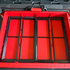 May include: A red and black tool storage drawer with an open front, featuring four vertical compartments separated by black plastic dividers. The drawer is part of a larger modular storage system with a black top handle and red latch mechanism.