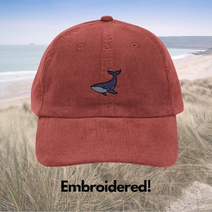 May include: A rust-colored corduroy baseball cap with a navy blue whale embroidery. The cap features a curved brim and a top button. The text "Embroidered!" is printed at the bottom of the image.
