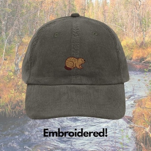 May include: An olive green corduroy baseball cap with an embroidered beaver design in brown shades. The cap has a curved peak and an adjustable strap. The text "Embroidered!" is displayed at the bottom of the image.