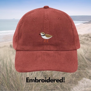 May include: A brick-red corduroy baseball cap featuring an embroidered bird. The bird is detailed in brown, white, and black, with orange legs. The text "Embroidered!" is at the bottom of the image.
