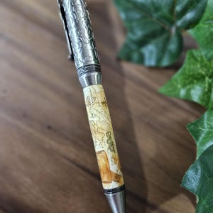 May include: A silver pen with a detailed engraved design on the top portion. The pen has a cream-colored barrel with a brown and gold map-like design. The pen tip is silver.