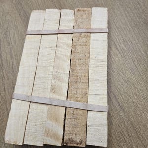 Op de afbeelding: Een klein, rechthoekig houten voorwerp bestaande uit verschillende lichtgekleurde houten latten die bij elkaar worden gehouden door twee bruine elastiekjes. De houtnerf is zichtbaar en sommige latten zijn donkerder dan andere.