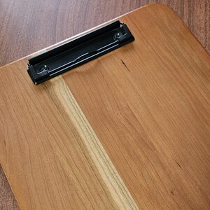 May include: A wooden clipboard with a black metal clip. The clipboard is made of light brown wood with visible grain and a smooth finish. The clip is attached with two screws.