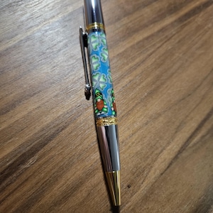 Könnte beinhalten: Ein silberner Stift mit einem blauen und grünen gemusterten Schaft mit einem Kleeblatt-Design und einer goldenen Verzierung. Der Stift hat einen silbernen Clip und eine goldene Spitze.