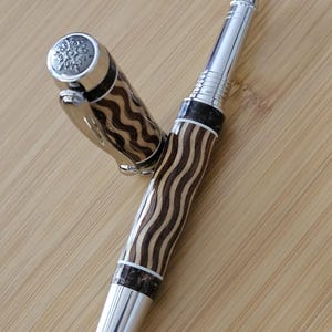 May include: A handcrafted rollerball pen with a silver-tone tip and accents. The pen's barrel features a wavy pattern of light and dark brown wood. The cap has a decorative silver-tone top. The pen is lying on a light brown wooden surface.