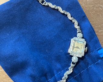 Könnte beinhalten: Eine silberne Uhr mit quadratischem Zifferblatt und einem mit Diamanten besetzten Band. Die Uhr liegt auf einem blauen Samtbeutel.