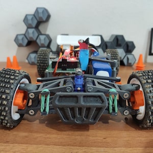 Puede incluir: Un coche de control remoto impreso en 3D negro y gris con ruedas blancas y detalles naranjas. El coche tiene un parachoques delantero negro y una carrocería azul y negra. El coche está sobre una superficie de madera.