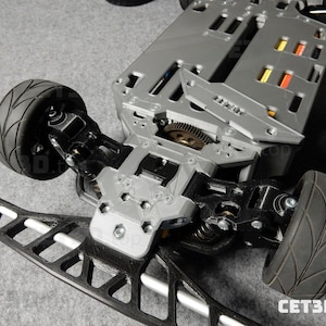 May include: A detailed view of a gray and black remote control car chassis. The chassis features black tires, suspension components, and a complex internal mechanism. The front bumper is black and white. The image is taken from a low angle.