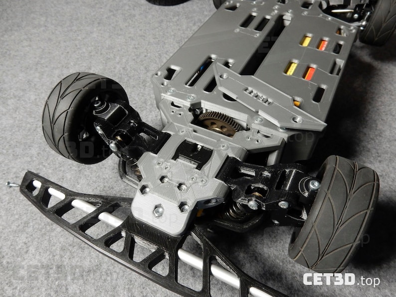May include: A detailed view of a gray and black remote control car chassis. The chassis features black tires, suspension components, and a complex internal mechanism. The front bumper is black and white. The image is taken from a low angle.