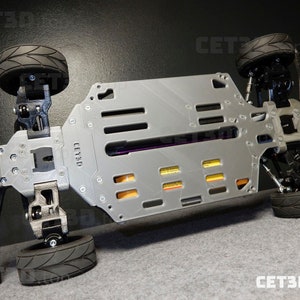 May include: A gray and black remote control car chassis with four black tires. The chassis has a rectangular design with multiple cutouts and the text "CET3D" is visible. The tires have a tread pattern.
