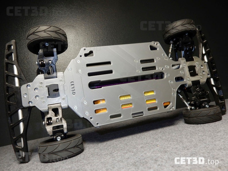 May include: A gray and black remote control car chassis with four black tires. The chassis has a rectangular design with multiple cutouts and the text "CET3D" is visible. The tires have a tread pattern.