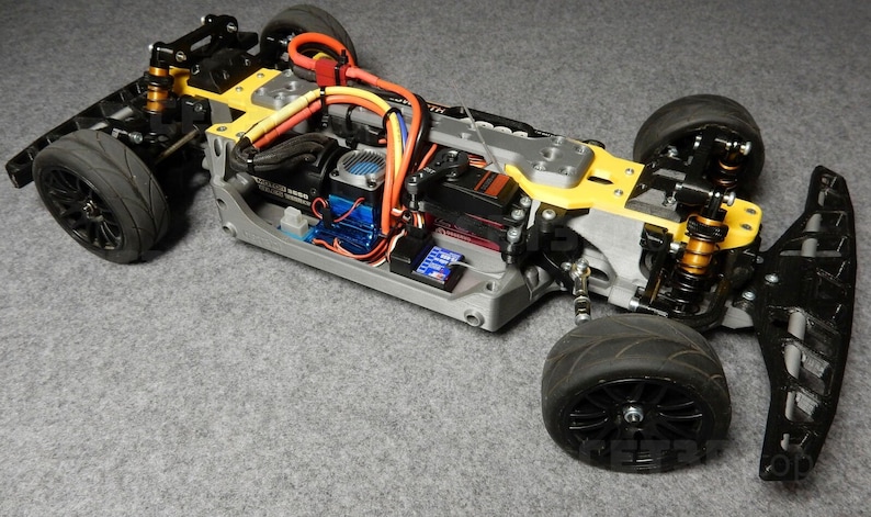 May include: A detailed view of a gray and yellow remote-controlled car chassis. The car features black tires, orange and yellow wiring, and a blue motor. The chassis is on a gray surface.