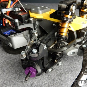 May include: Close-up of a 3D-printed RC car chassis. The chassis features black, gray, and yellow components, including suspension, shocks, and a purple bolt. Wires and electronic components are visible.