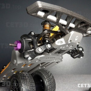 May include: A detailed view of a 3D-printed RC car chassis. The chassis is primarily gray, with black, purple, and yellow accents. The tires are black with a tread pattern. The image is taken from a low angle, showcasing the car's suspension and undercarriage.