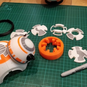 Könnte beinhalten: Ein weißer und orangefarbener 3D-gedruckter BB-8-Droide mit einem schwarzen Ring, einem Stapel orangefarbener Ringe und weißen und grauen Teilen zum Zusammenbauen. Ein Klebstoff und ein schwarzer Stift sind ebenfalls enthalten.