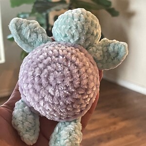 Könnte beinhalten: Ein gehäkeltes Schildkrötenspielzeug mit einer hellblauen Schale und einem violetten Körper. Das Spielzeug ist aus weichem, flauschigem Garn gefertigt.